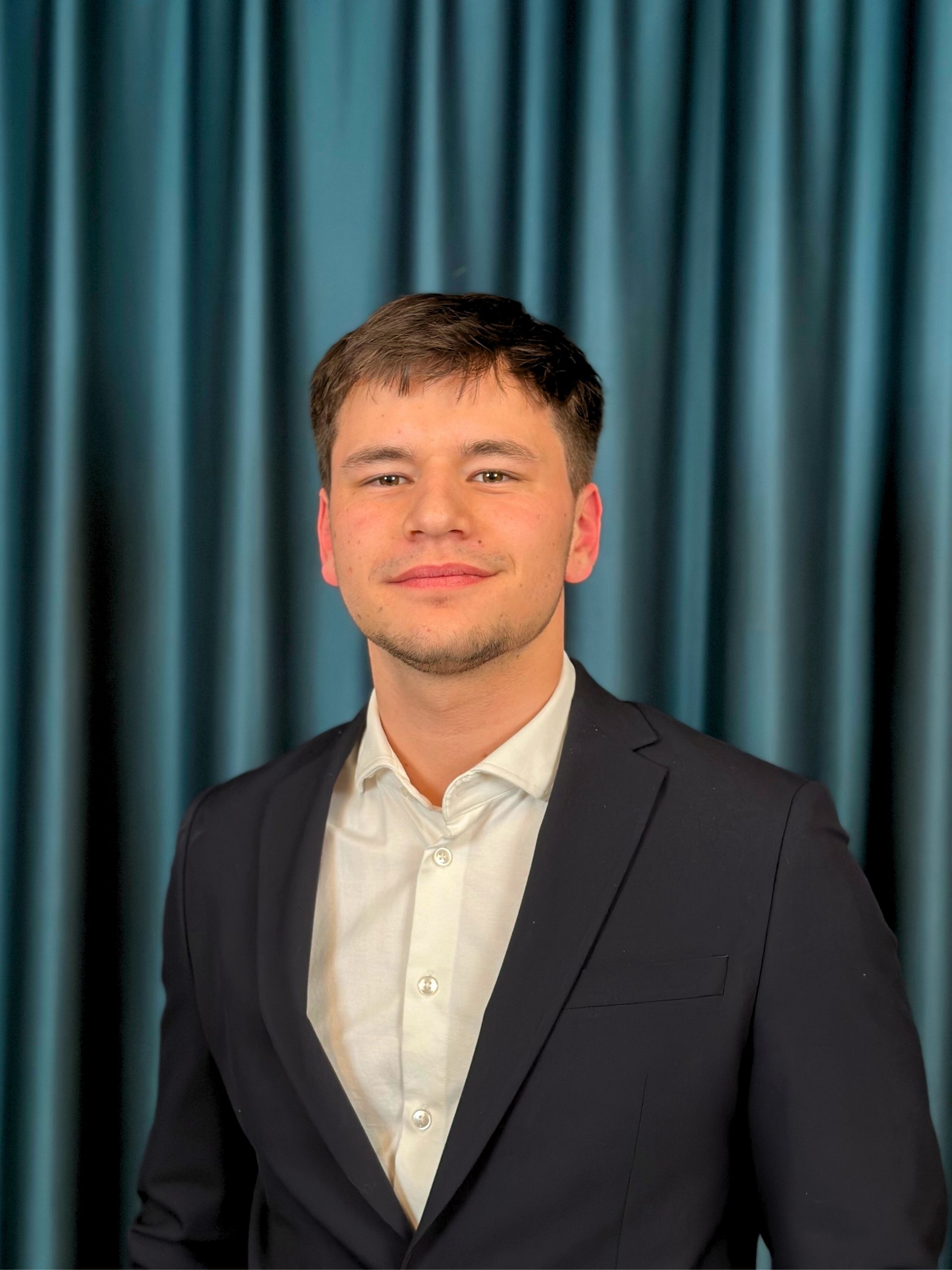 Young professional man with short hair, wearing a black suit and a white shirt, posing confidently in front of a dark teal curtain background.