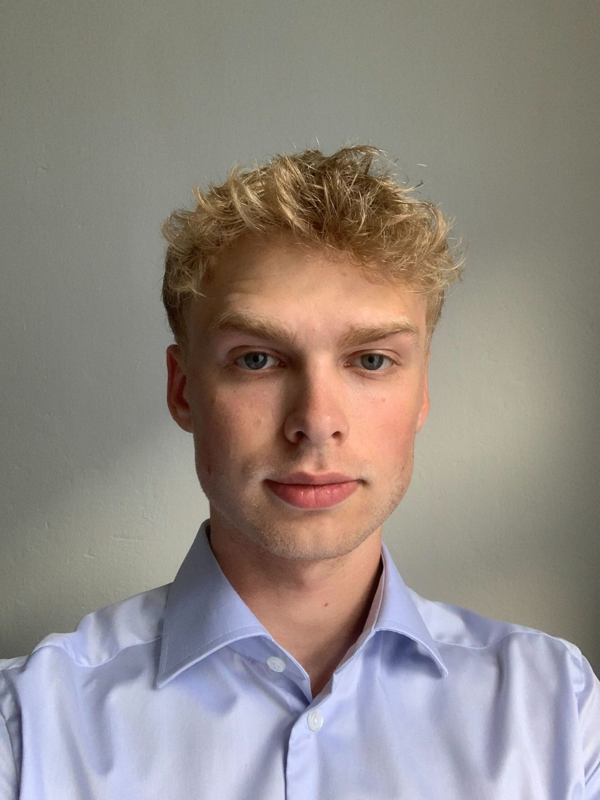 Headshot of a young man with blond hair, wearing a light blue shirt, looking at the camera with a neutral expression, against a grey background.