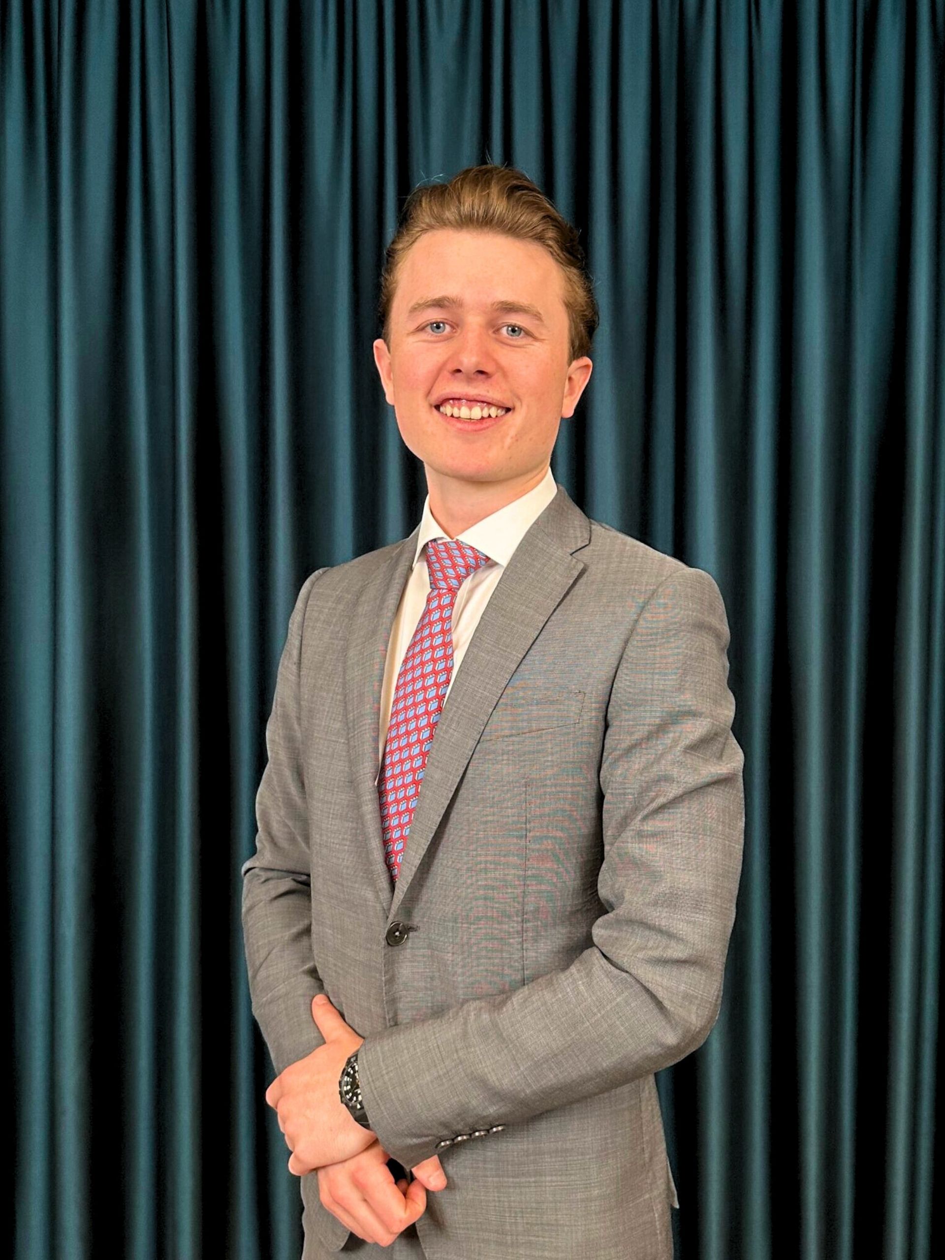 Confident young businessman in grey suit and with a red patterned tie standing with crossed arms in front of a teal curtain background.