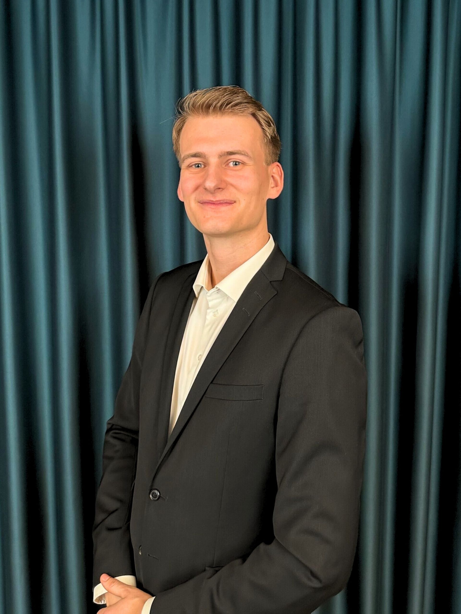 Young professional blonde man with a neat hairstyle, wearing a black suit and a white shirt, posing confidently in front of a dark teal curtain background.