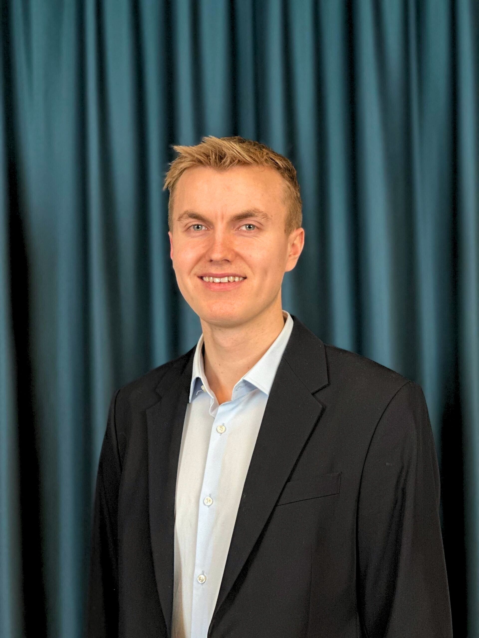 Professional headshot of a smiling young man with blonde hair wearing glasses, a light blue shirt, and a black blazer, with a teal curtain background.