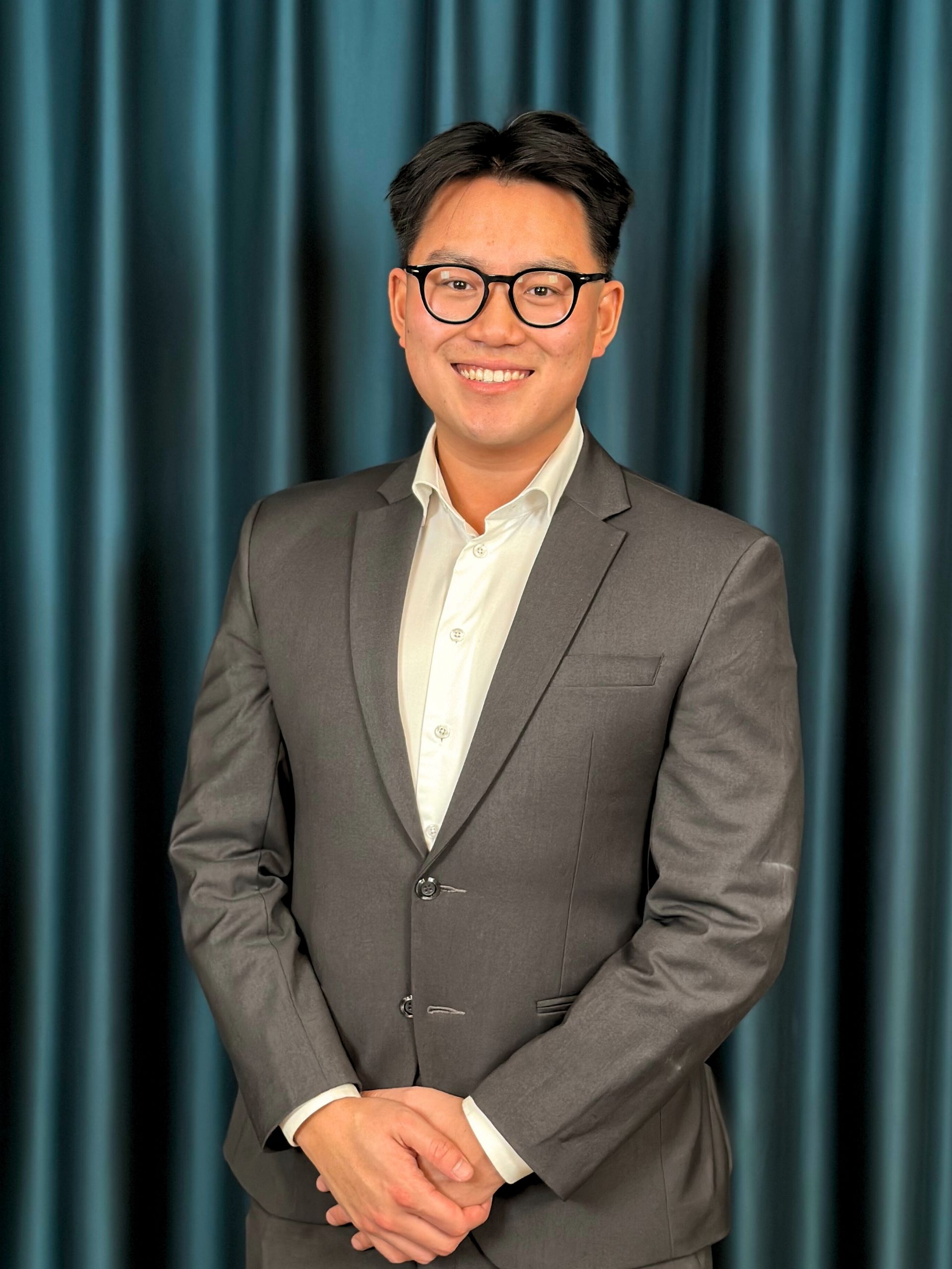 Young professional man with black glasses, wearing a grey suit and a white shirt, posing with a friendly smile in front of a dark teal curtain background.