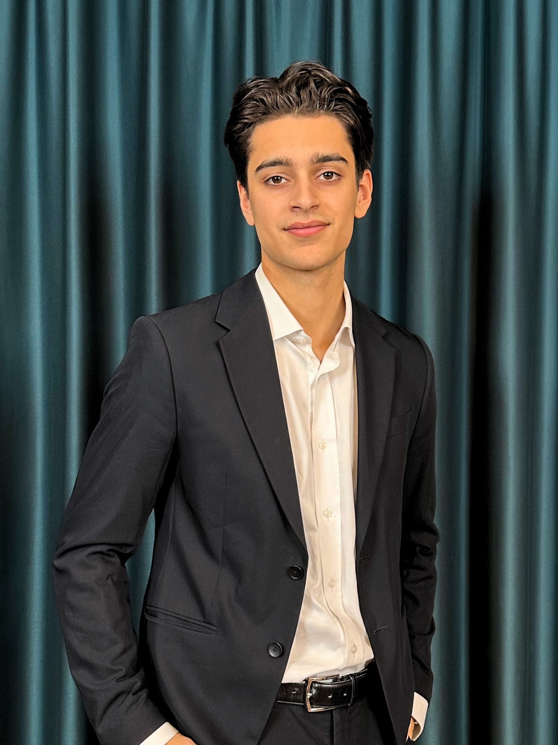 Young professional man with a neat hairstyle, wearing a black suit and a white shirt, posing confidently in front of a dark teal curtain background.