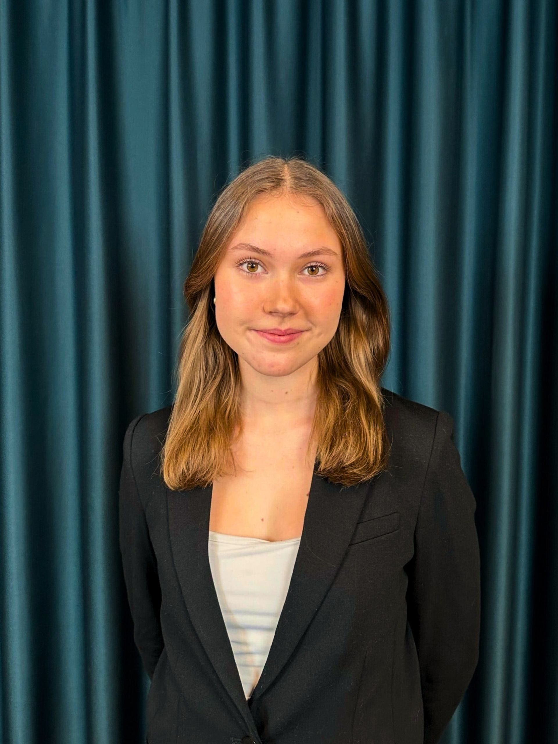 Professional headshot of a young woman with medium lenght hair, wearing a black blazer and white top, against a teal curtain backdrop.