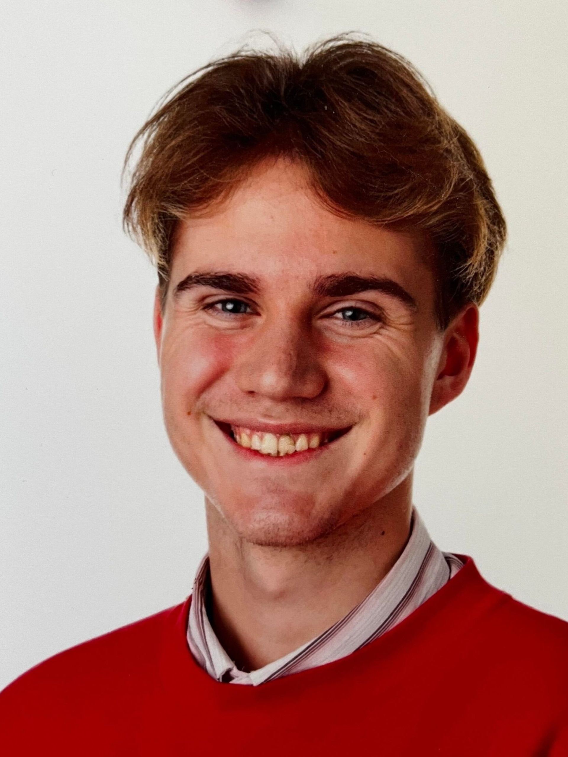 Portrait of a cheerful young man with light brown hair, wearing a red sweater and collared shirt, smiling in front of a white background.