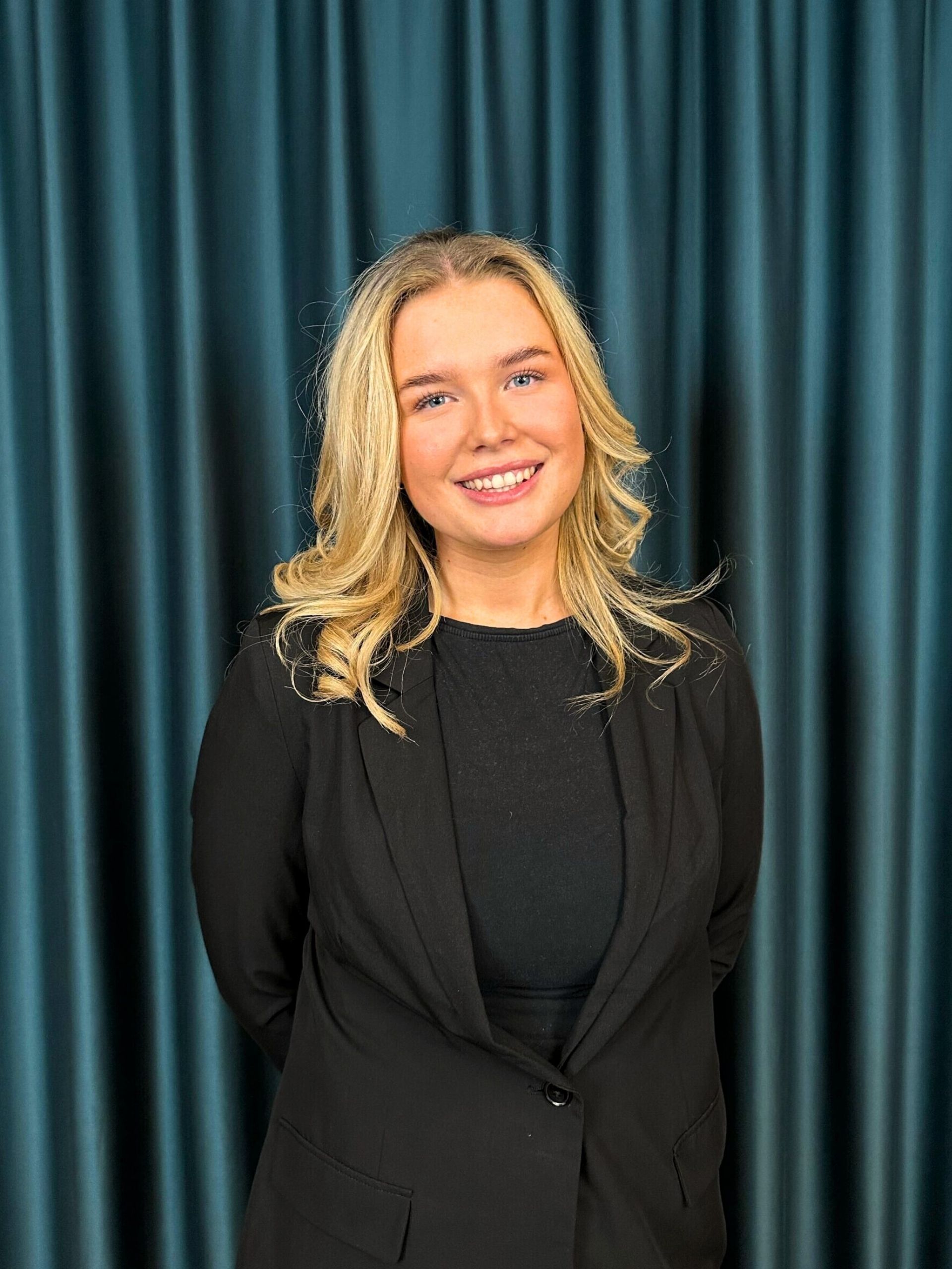 Professional headshot of a smiling blonde woman in a black blazer and black top, against a teal curtain backdrop.