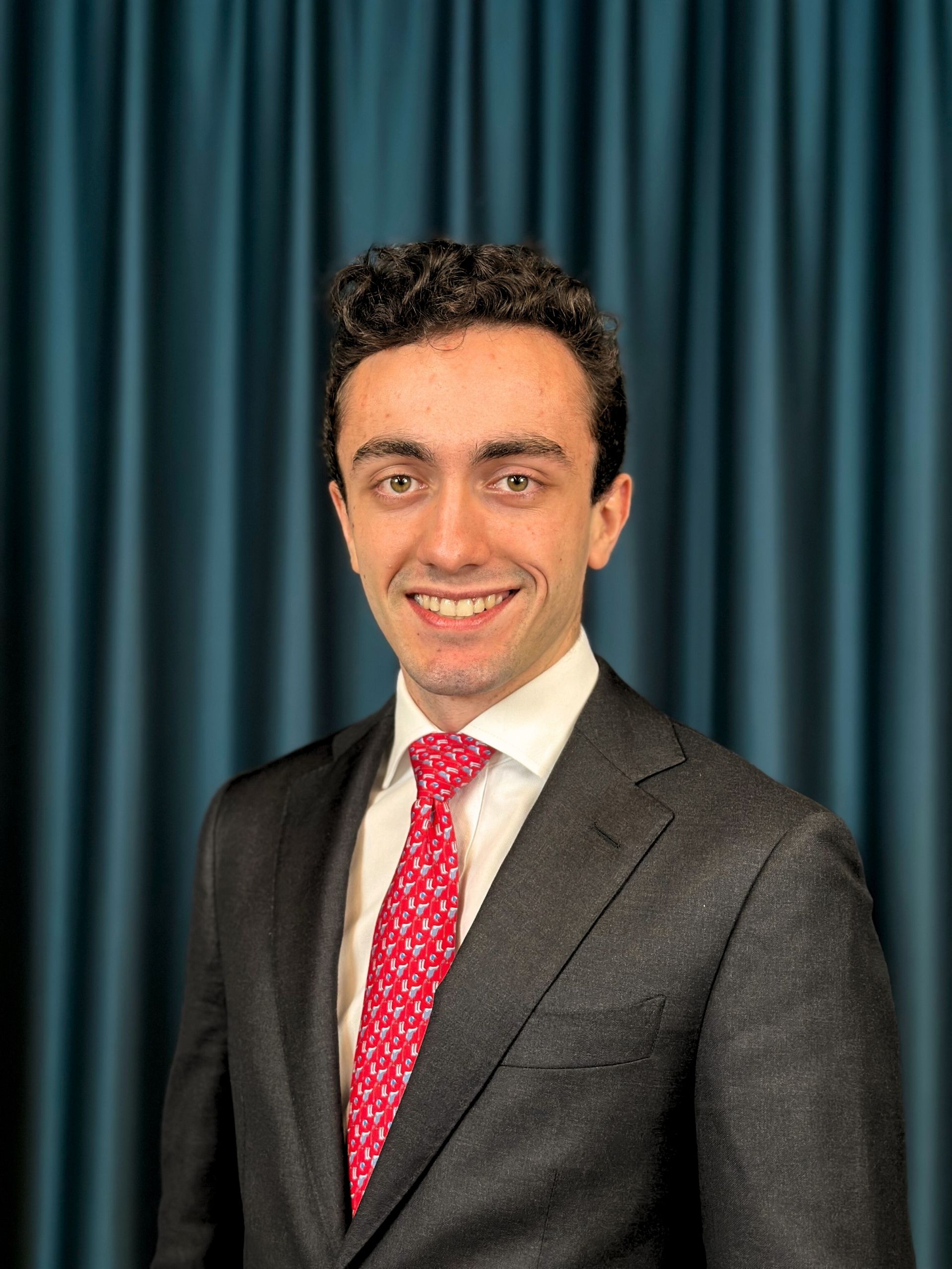 Professional headshot of a smiling man with short curly hair in a grey blazer, white shirt and a red tie against a teal curtain backdrop.