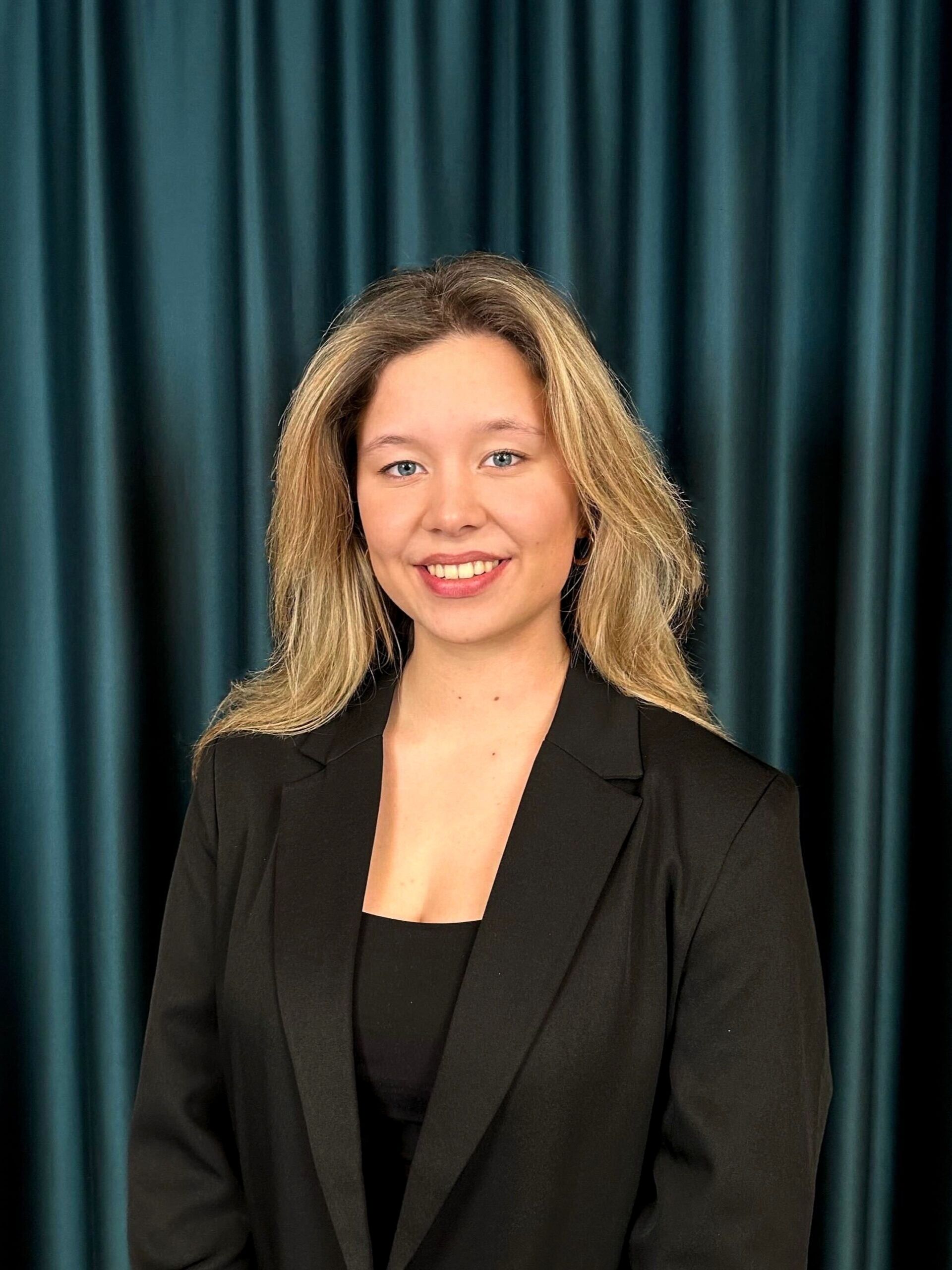 Professional headshot of a smiling blonde woman in a black blazer and a black top, against a teal curtain backdrop.