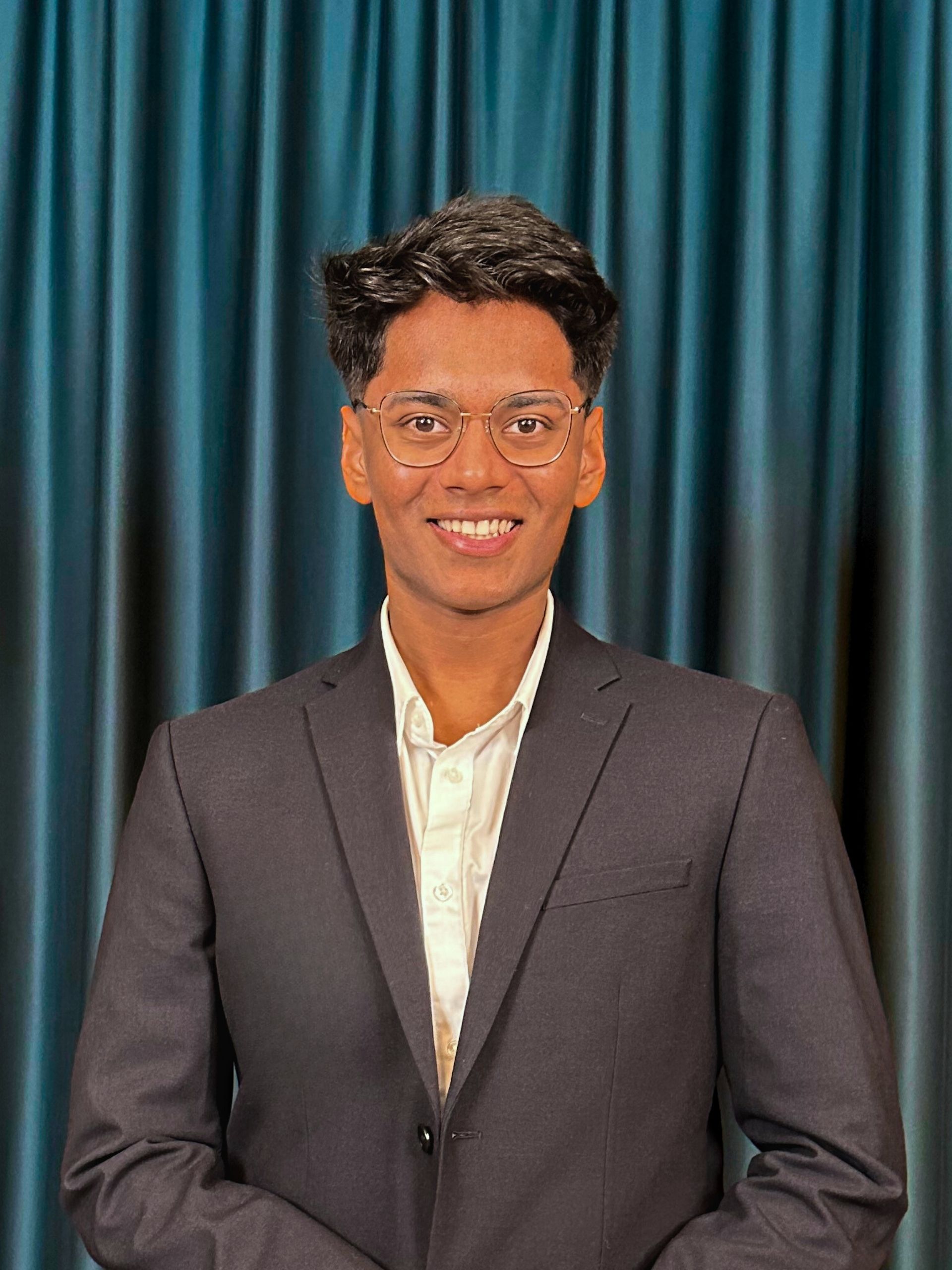 Professional headshot of a smiling young man with black hair wearing glasses, a white shirt, and a black blazer, with a teal curtain background.