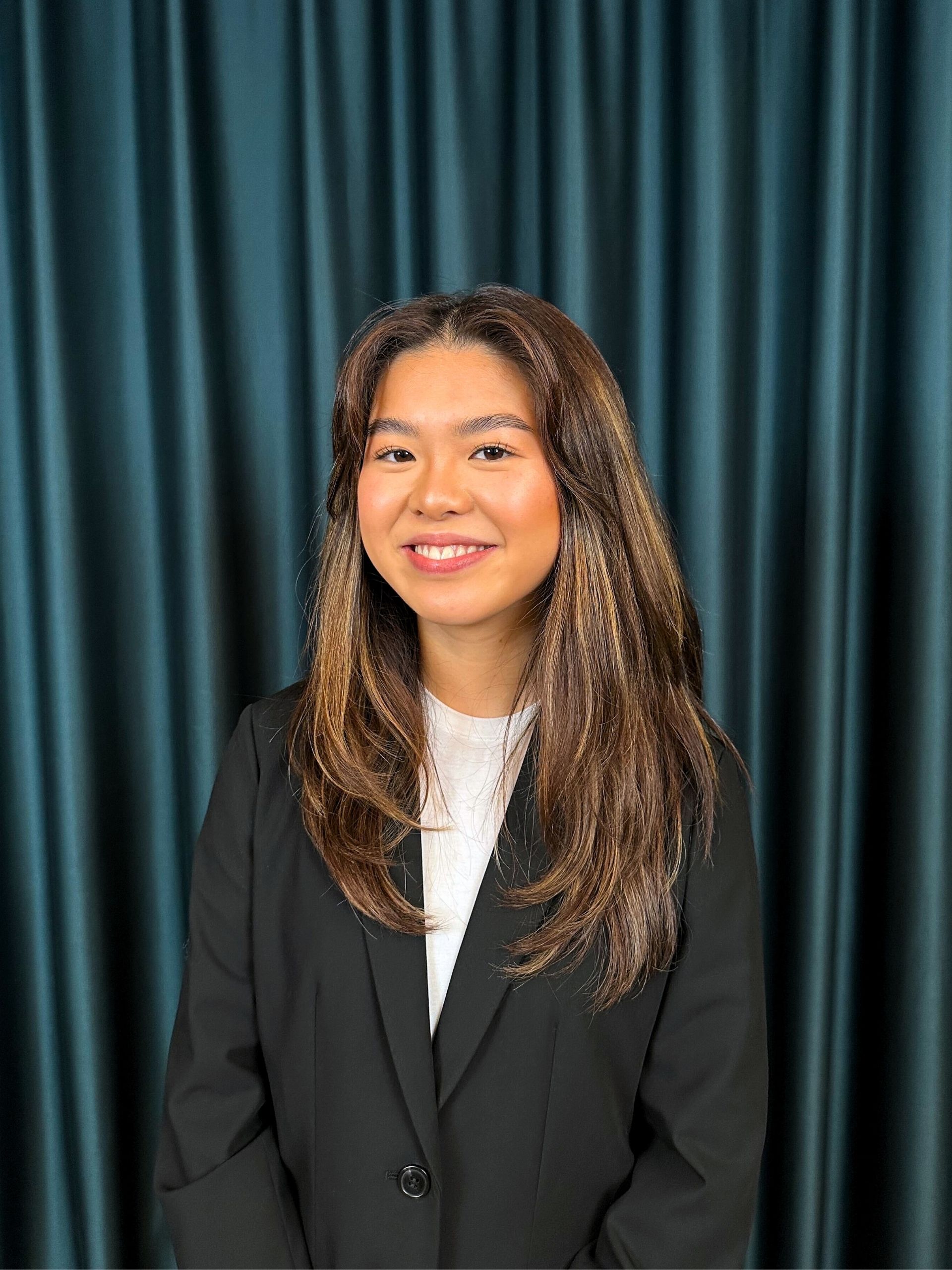 Young professional woman with long brown hair, wearing a black suit and a white top, posing with a friendly smile in front of a dark teal curtain background.