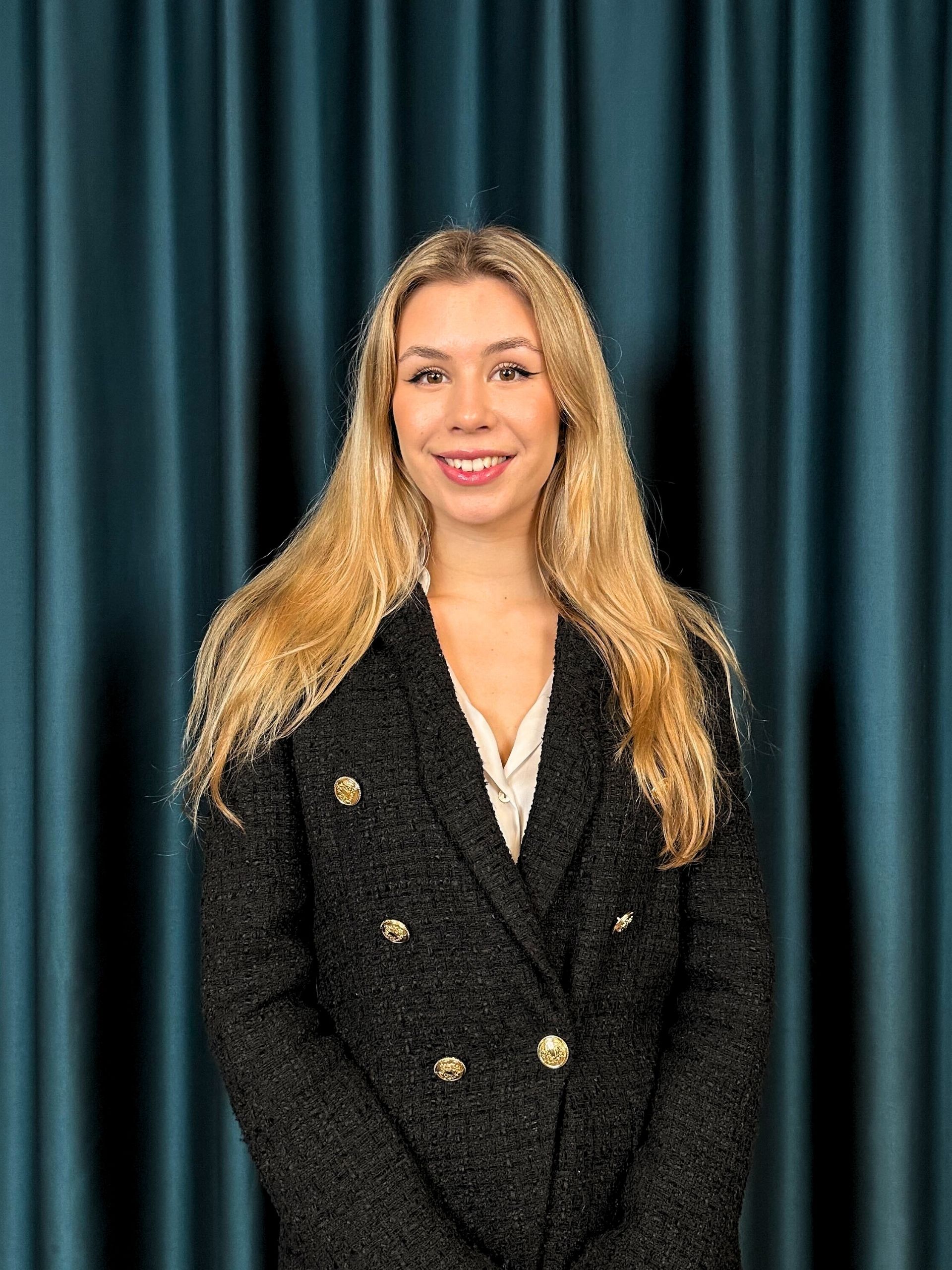 Professional headshot of a smiling blonde woman in a black tweed blazer and a cream top, against a teal curtain backdrop.