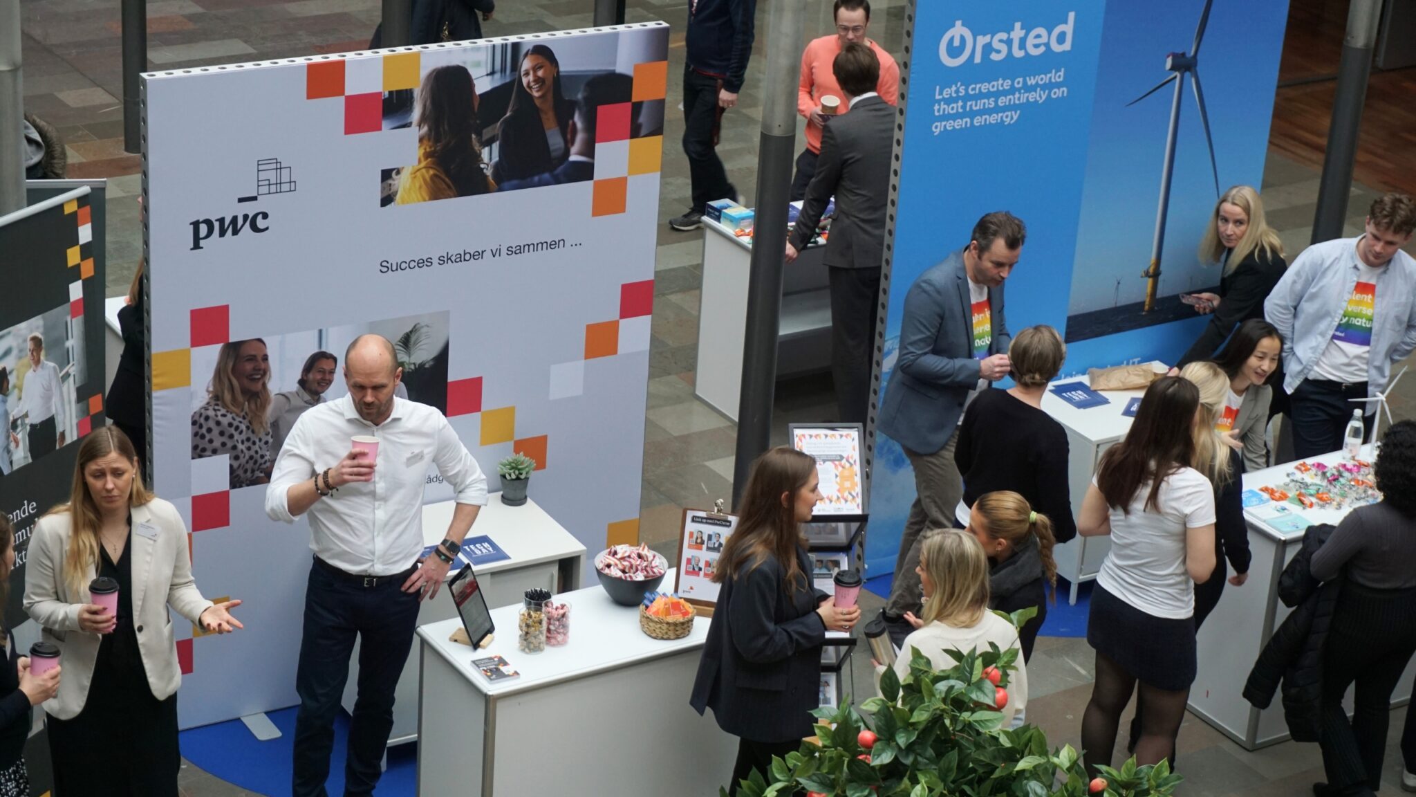 An aerial view of a bustling exhibition area at CBS Tech Day. Attendees interact with representatives at various company booths, like PwC and Ørsted, which are prominently displayed with large banners. The PwC booth features a message of collaboration, while Ørsted emphasizes green energy with an image of a wind turbine. The scene is dynamic, with individuals engaged in conversation, exchanging information, and some attendees holding coffee cups, all contributing to the networking vibe of the event.