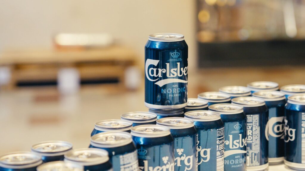 A close-up image of Carlsberg non-alcoholic beer cans arranged on a table, with one can standing prominently in focus against a blurred background. The cans display the Carlsberg Nordic Pilsner branding, indicating a beverage choice offered to attendees at the event. The arrangement suggests a casual and inclusive setting, catering to diverse preferences and promoting a responsible drinking environment.