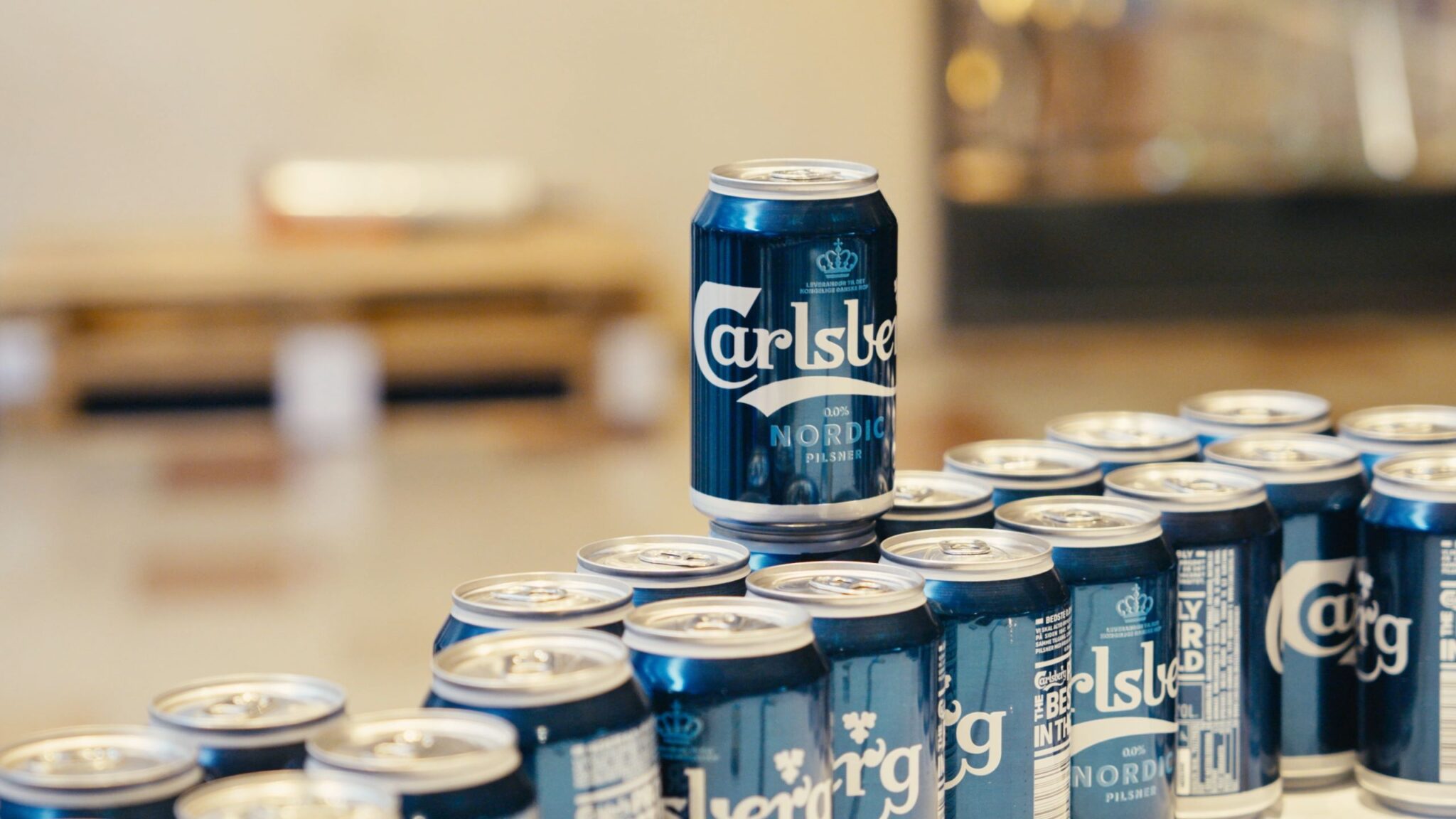 A close-up image of Carlsberg non-alcoholic beer cans arranged on a table, with one can standing prominently in focus against a blurred background. The cans display the Carlsberg Nordic Pilsner branding, indicating a beverage choice offered to attendees at the event. The arrangement suggests a casual and inclusive setting, catering to diverse preferences and promoting a responsible drinking environment.