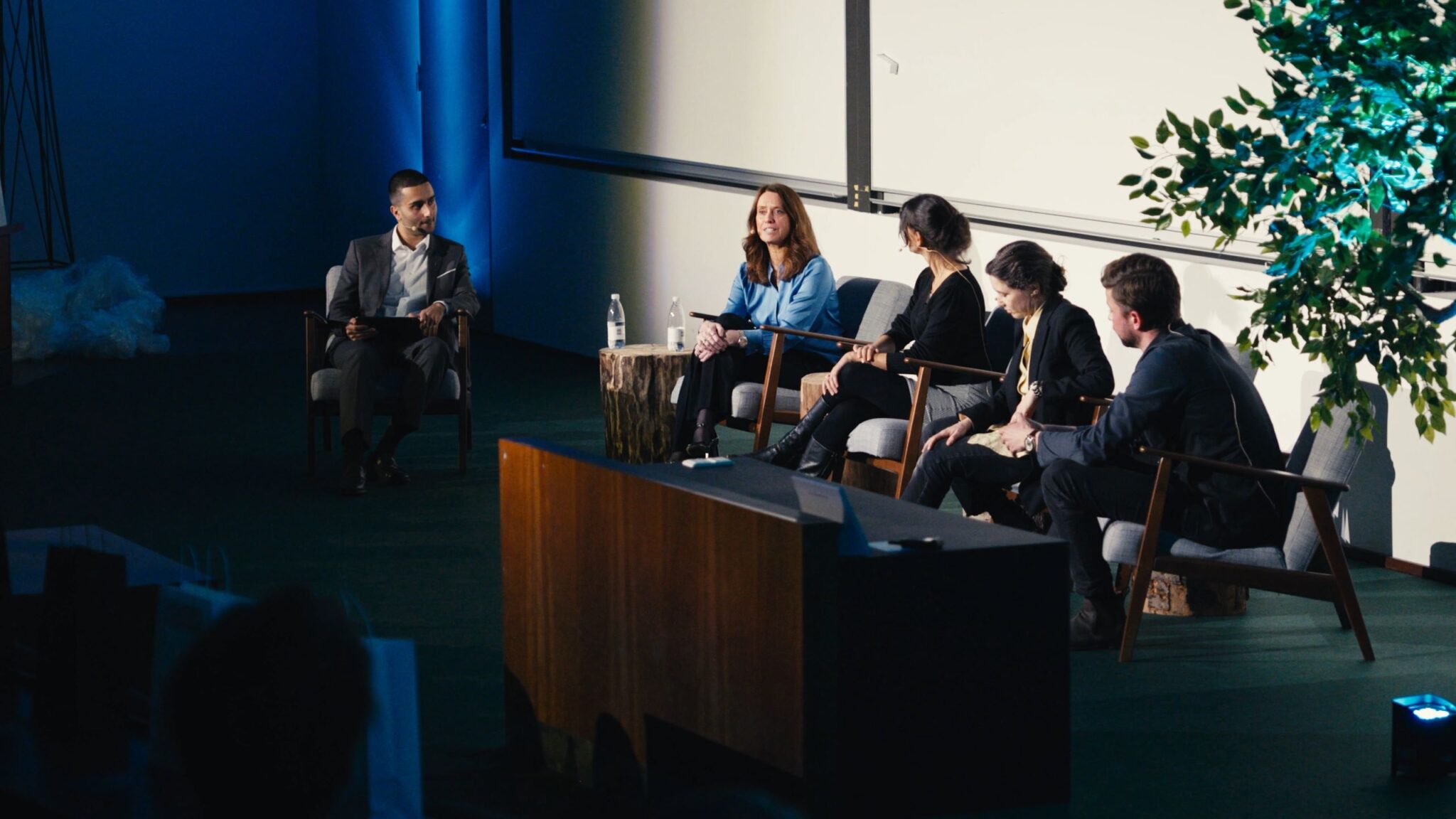 Close-up view of a debate panel at CBS Tech Day, featuring a moderator and four panelists engaged in a focused discussion. The panelists, dressed in professional attire, are seated in a row on stage with natural elements like a tree adding a touch of greenery to the setting. The spotlight illuminates the panel against a background of soft blue lighting, creating a serious yet inviting atmosphere for the exchange of ideas on technology.