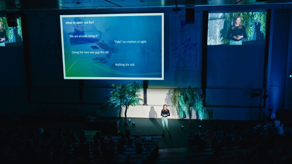 The auditorium at CBS Tech Day is cast in soft blue tones, focusing attention on the keynote speaker concluding the event. The speaker is displayed on a large screen alongside a presentation slide about best practices and warnings in technology innovation. The slide reads, "What to watch out for?" with bullet points such as "We are already doing it" and "Walking the talk". The audience, seen in silhouette, watches attentively from their seats, surrounded by lush greenery that decorates the stage, enhancing the event's ambiance.