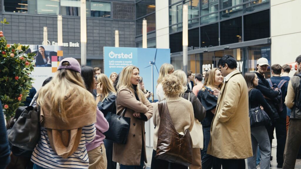 Energetic networking scene at CBS Tech Day where students mingle with industry professionals. The indoor setting is abuzz with conversation as groups engage with representatives from top tech companies. In the foreground, we see attendees in casual and business casual attire, exchanging ideas and contact details. Promotional banners from companies like Ørsted, featuring a wind turbine image, suggest a focus on sustainable technology. The atmosphere is informal and collaborative, with potential opportunities for internships and careers being discussed.