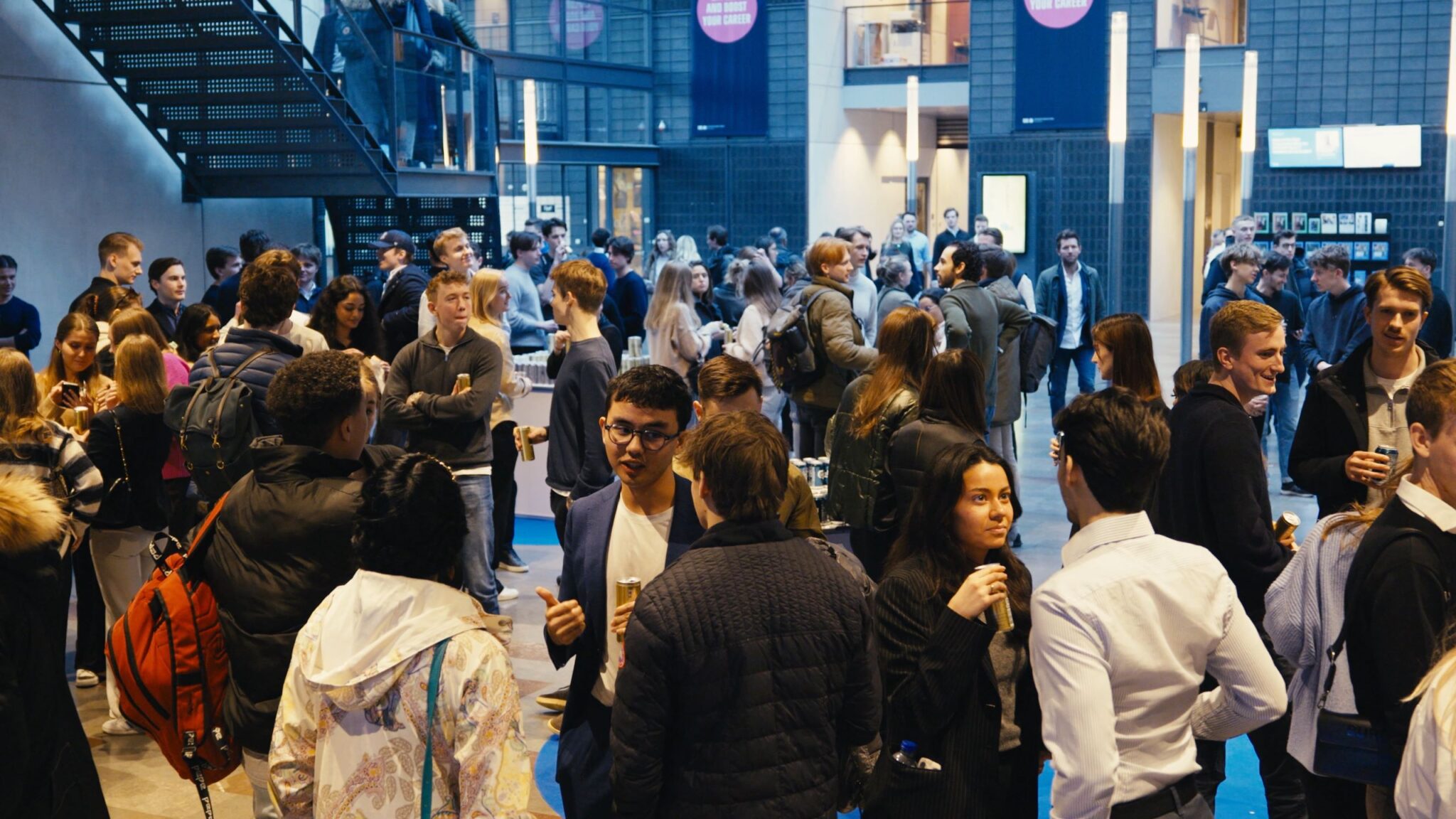 A lively networking break at CBS Tech Day, with students and young professionals engaging in conversations. The hall is filled with a diverse group of individuals, some standing and chatting, others walking through, all amidst a backdrop of modern architecture. The atmosphere is casual and interactive, with many participants holding drinks, indicative of a relaxed environment conducive to building connections between debate panels.