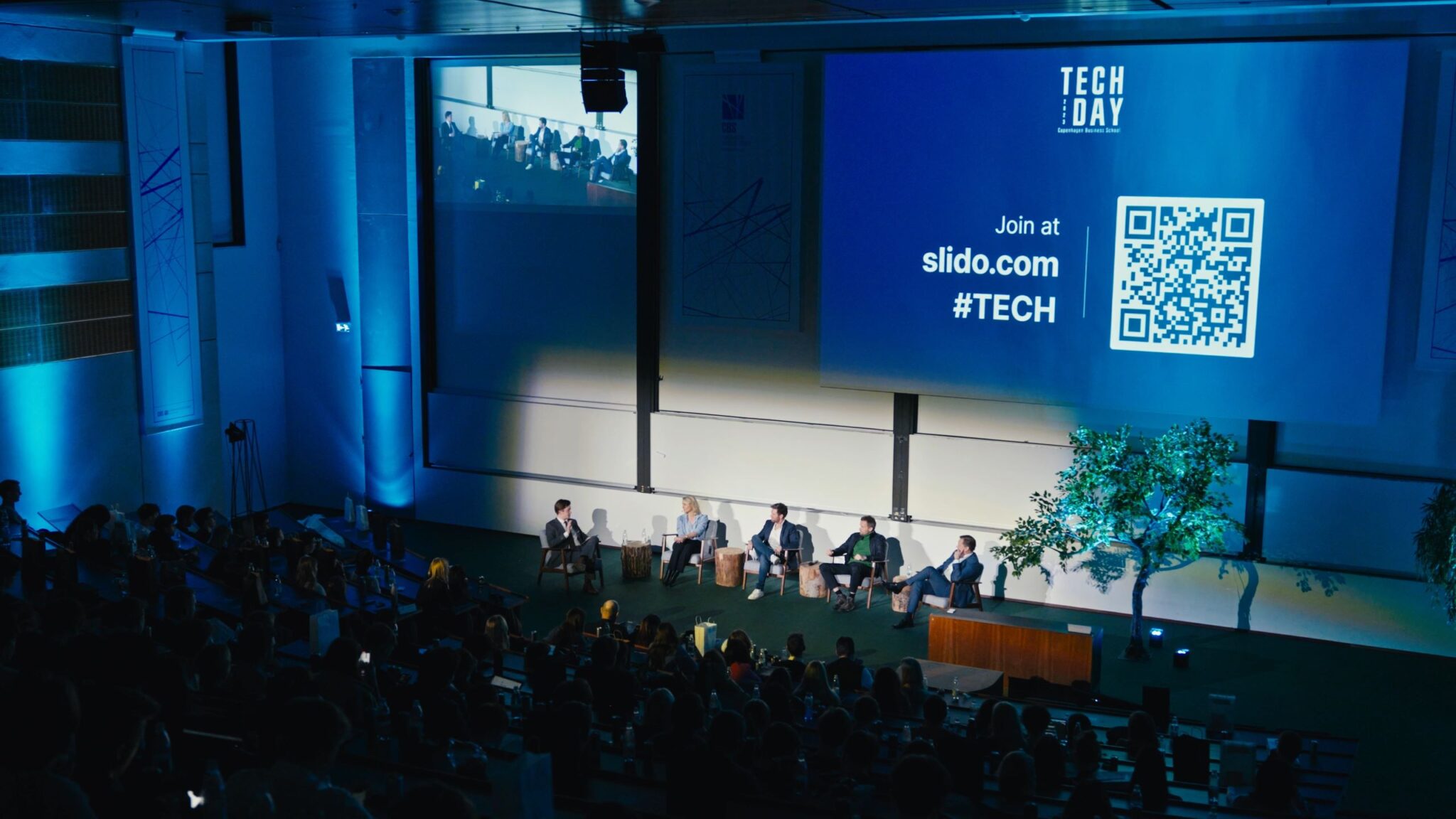 A debate panel at the CBS Tech Day with a moderator and four tech leaders seated on stage, engaging in discussion. The auditorium is dimly lit, highlighting the stage with a large screen displaying "TECH DAY" branding and a QR code for joining the conversation on Slido with the hashtag #TECH. The audience is in shadow, directing the focus to the panelists who appear to be sharing insights on the latest tech issues.