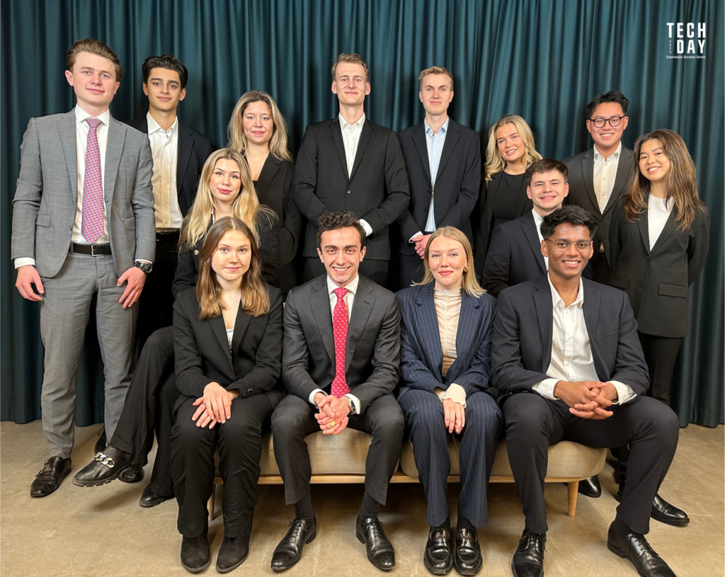 Group of professionally dressed young adults smiling for a photo at Tech Day event, with a teal curtain background and the event's logo visible.