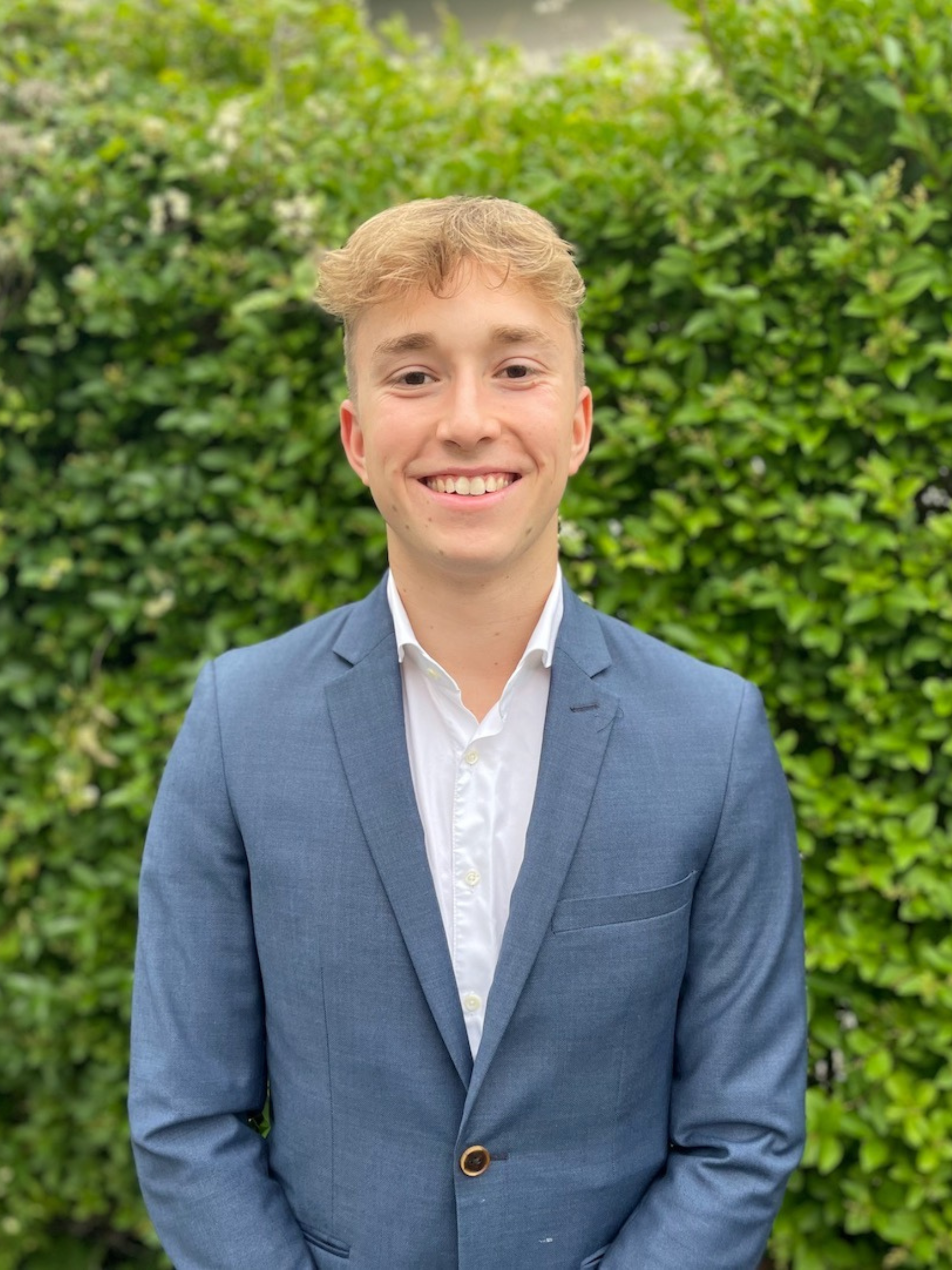 Cheerful young man with short blond hair smiling for a professional headshot in a blue blazer and white shirt, with a lush green hedge in the background.