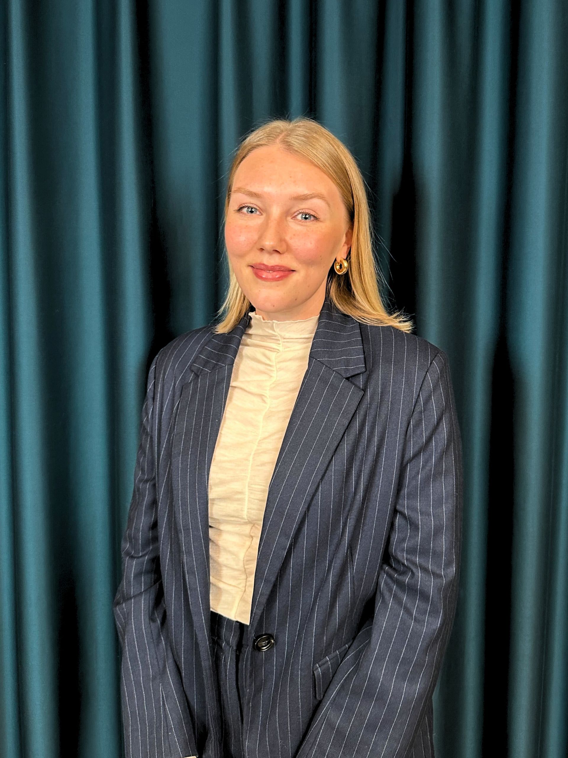 Professional headshot of a smiling blonde woman in a pinstriped blazer and beige blouse, with a teal curtain background.