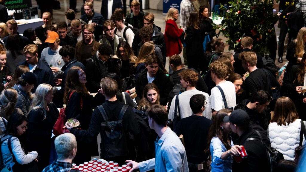 A large group of people mingling and enjoying drinks and food at a tech conference after-debate event. The crowd is gathered around tables with free food and drinks, creating a lively and bustling atmosphere.