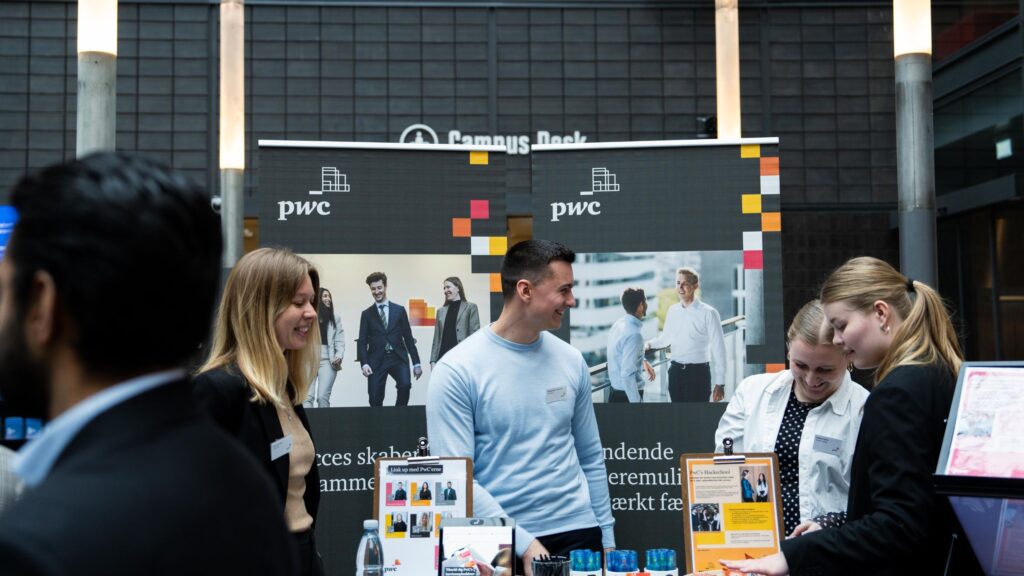 Young professionals and students engaging at the PwC booth during a tech conference. The booth features PwC branding and images of diverse professionals. The attendees are smiling and discussing opportunities, creating a lively and positive atmosphere.