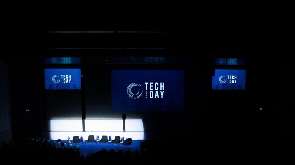 An empty debate venue at Tech Day, featuring a stage with several chairs and a large screen displaying the Tech Day logo. The room is dimly lit, creating a sense of anticipation for the upcoming panel discussion.