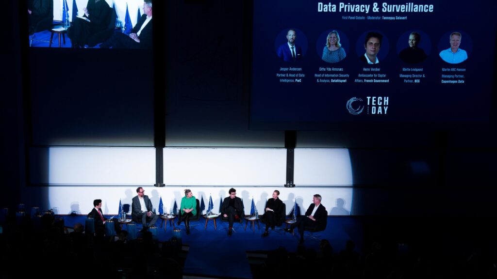 A panel of tech industry leaders seated on stage during a Tech Day debate on data privacy and surveillance. A large screen above them displays their names, titles, and event details. The audience watches as the experts share insights and engage in discussion.