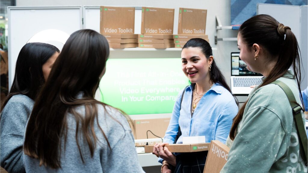 A group of students chatting with a representative from a partner company at a tech conference. They are smiling and holding video toolkit boxes. A backdrop with product information and a laptop is visible in the background.