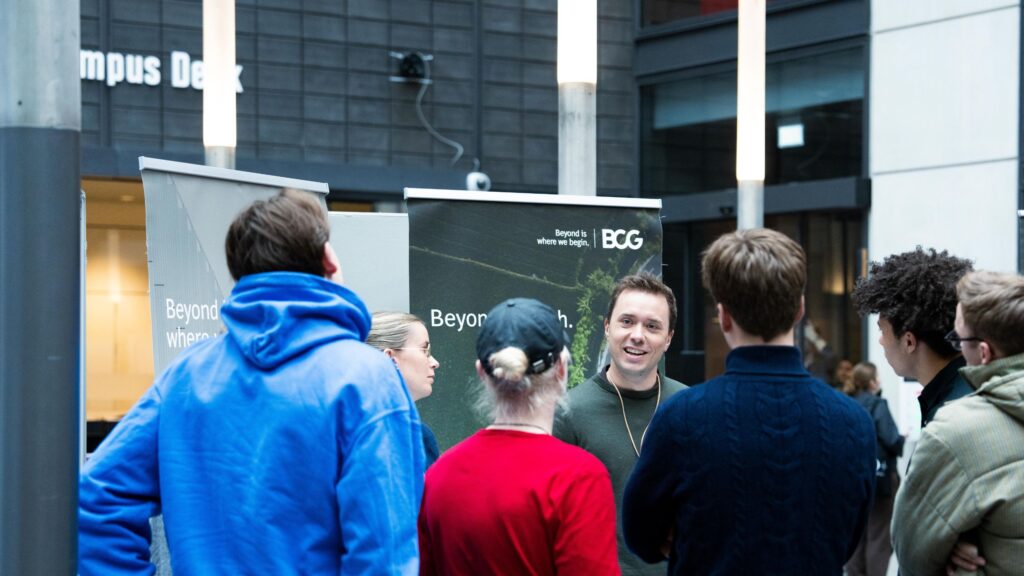 A group of students engaging in a conversation with a representative from BCG at a tech conference. The discussion is taking place in front of a BCG banner that reads "Beyond is where we begin," highlighting the company's innovative approach. **Alt Text:** Students talking with a representative from BCG at a tech conference. The conversation is happening in front of a BCG banner with the slogan "Beyond is where we begin." The representative and students are smiling and engaging in a lively discussion.