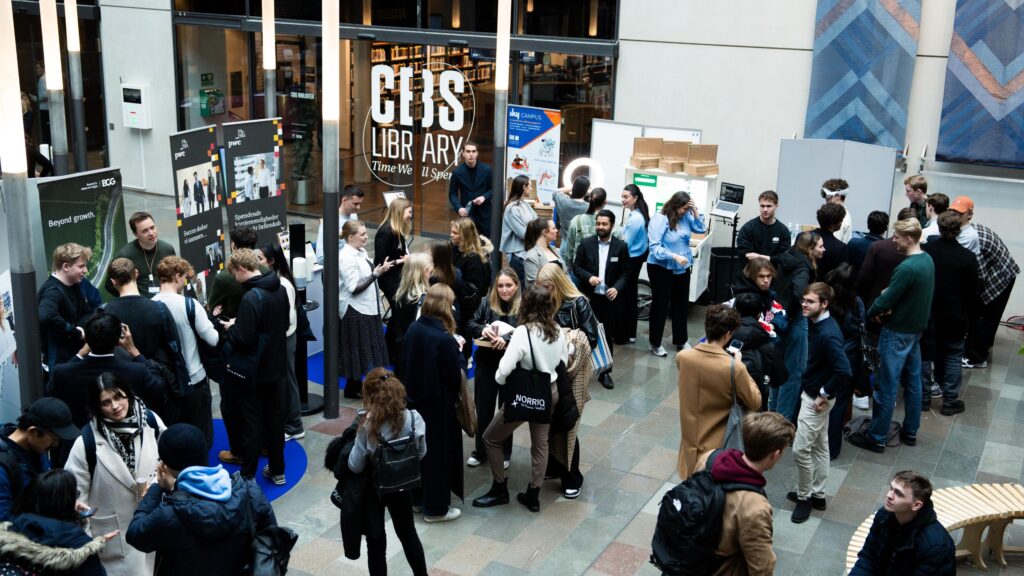 A busy exhibition hall at a tech conference where various companies are interacting with a large group of students. Multiple booths display company banners and products as attendees engage in discussions and explore opportunities.