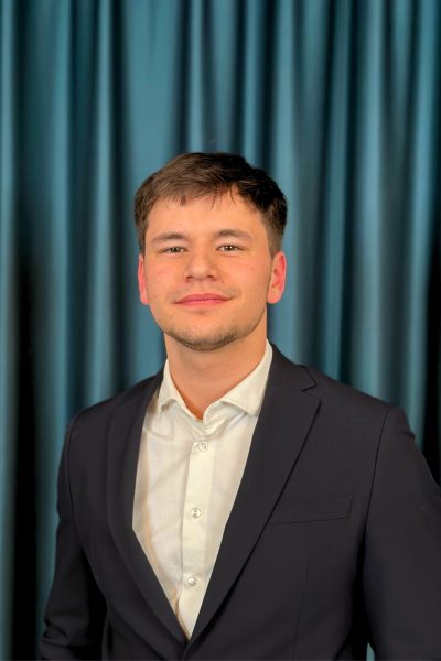 Young professional man with short hair, wearing a black suit and a white shirt, posing confidently in front of a dark teal curtain background.