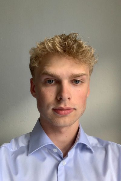Headshot of a young man with blond hair, wearing a light blue shirt, looking at the camera with a neutral expression, against a grey background.