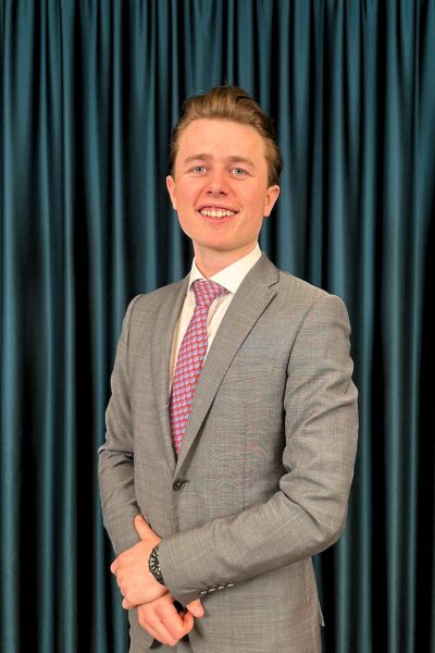 Confident young businessman in grey suit and with a red patterned tie standing with crossed arms in front of a teal curtain background.