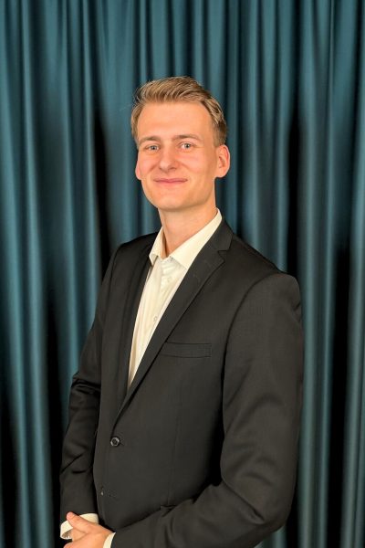 Young professional blonde man with a neat hairstyle, wearing a black suit and a white shirt, posing confidently in front of a dark teal curtain background.