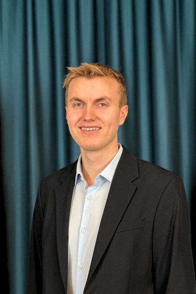 Professional headshot of a smiling young man with blonde hair wearing glasses, a light blue shirt, and a black blazer, with a teal curtain background.