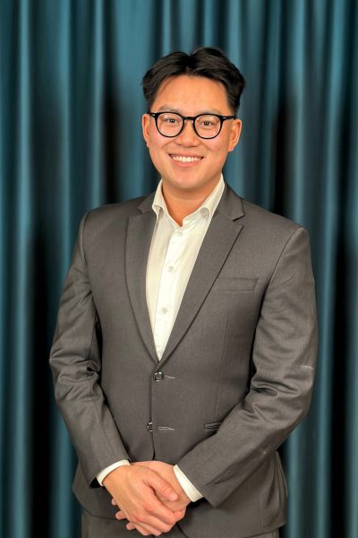Young professional man with black glasses, wearing a grey suit and a white shirt, posing with a friendly smile in front of a dark teal curtain background.