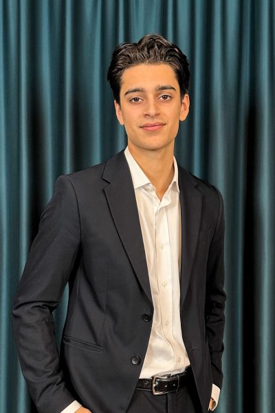 Young professional man with a neat hairstyle, wearing a black suit and a white shirt, posing confidently in front of a dark teal curtain background.