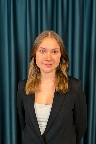 Professional headshot of a young woman with medium lenght hair, wearing a black blazer and white top, against a teal curtain backdrop.