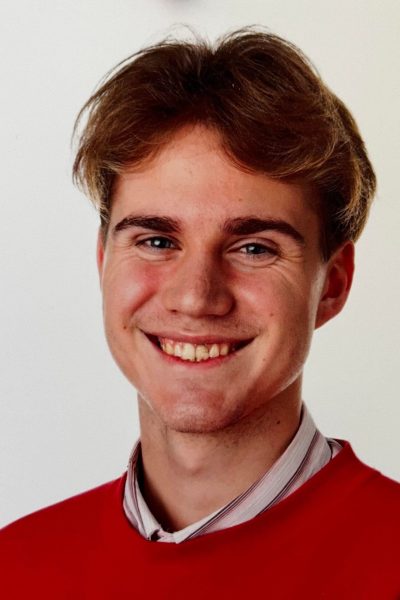 Portrait of a cheerful young man with light brown hair, wearing a red sweater and collared shirt, smiling in front of a white background.