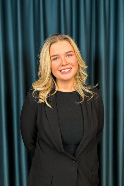 Professional headshot of a smiling blonde woman in a black blazer and black top, against a teal curtain backdrop.