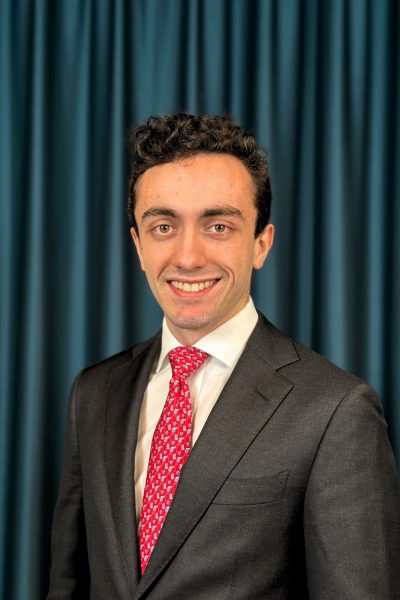 Professional headshot of a smiling man with short curly hair in a grey blazer, white shirt and a red tie against a teal curtain backdrop.
