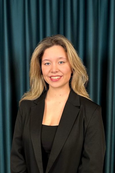 Professional headshot of a smiling blonde woman in a black blazer and a black top, against a teal curtain backdrop.