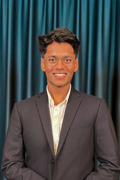 Professional headshot of a smiling young man with black hair wearing glasses, a white shirt, and a black blazer, with a teal curtain background.