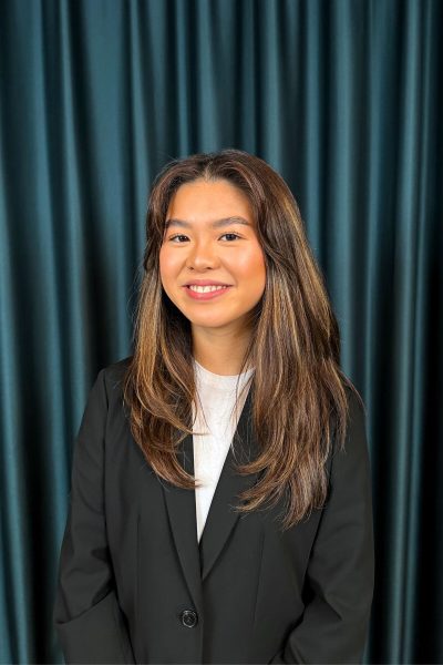Young professional woman with long brown hair, wearing a black suit and a white top, posing with a friendly smile in front of a dark teal curtain background.