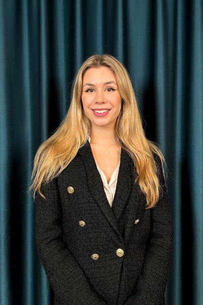 Professional headshot of a smiling blonde woman in a black tweed blazer and a cream top, against a teal curtain backdrop.