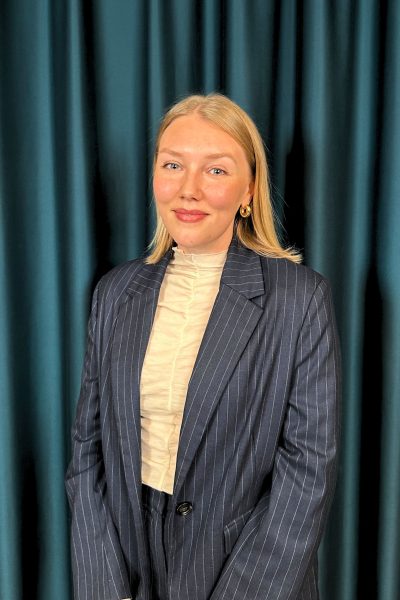 Professional headshot of a smiling blonde woman in a pinstriped blazer and beige blouse, with a teal curtain background.