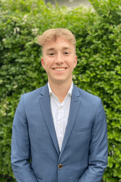 Cheerful young man with short blond hair smiling for a professional headshot in a blue blazer and white shirt, with a lush green hedge in the background.
